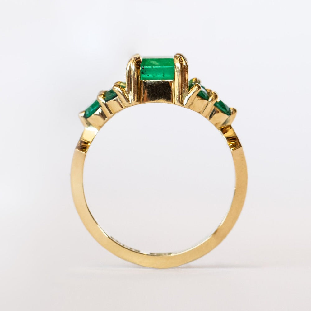 An emerald ring set of engagement ring and wedding band. Features an emerald cut emerald gemstone center flanked by six round emeralds, and a graduating size emerald band that contours to fit.