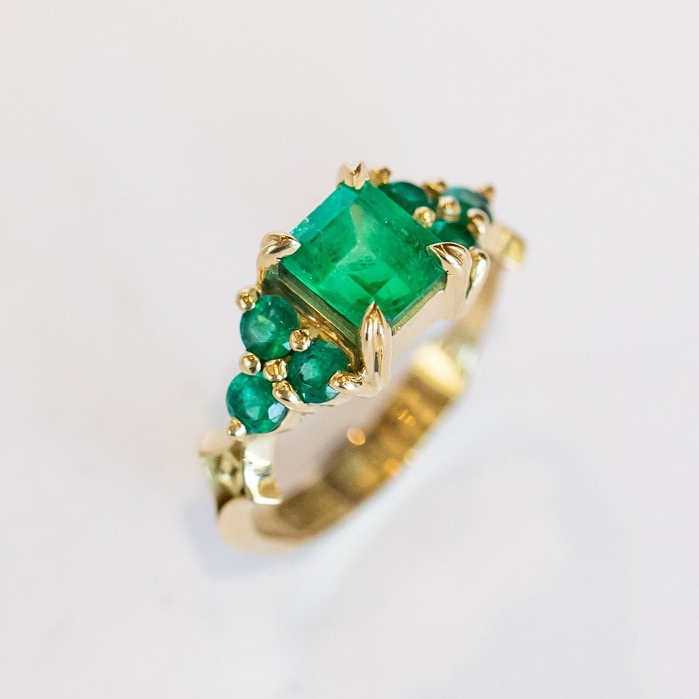 An emerald ring set of engagement ring and wedding band. Features an emerald cut emerald gemstone center flanked by six round emeralds, and a graduating size emerald band that contours to fit.