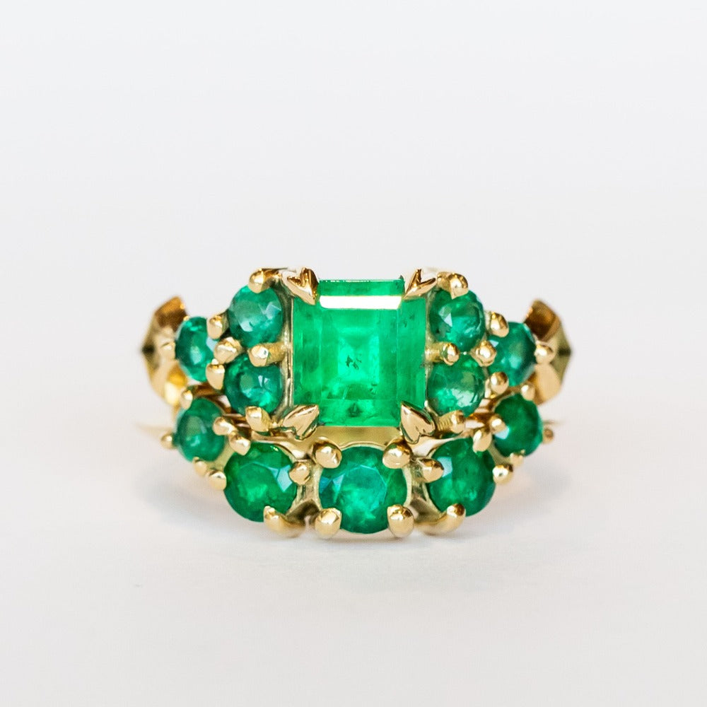 An emerald ring set of engagement ring and wedding band. Features an emerald cut emerald gemstone center flanked by six round emeralds, and a graduating size emerald band that contours to fit.