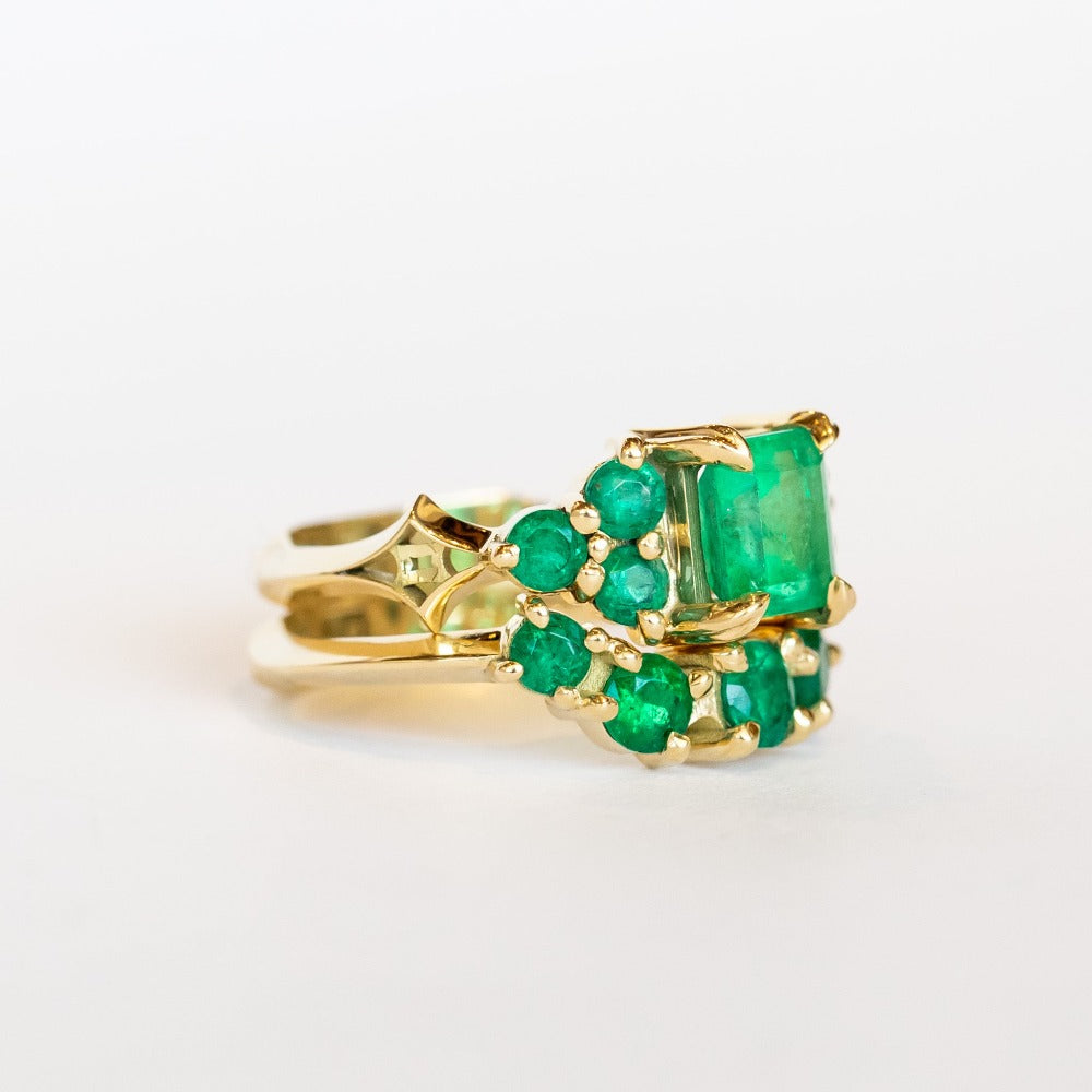 An emerald ring set of engagement ring and wedding band. Features an emerald cut emerald gemstone center flanked by six round emeralds, and a graduating size emerald band that contours to fit.