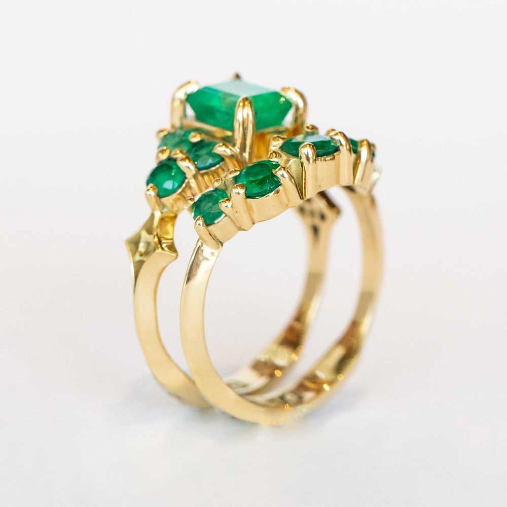 An emerald ring set of engagement ring and wedding band. Features an emerald cut emerald gemstone center flanked by six round emeralds, and a graduating size emerald band that contours to fit.