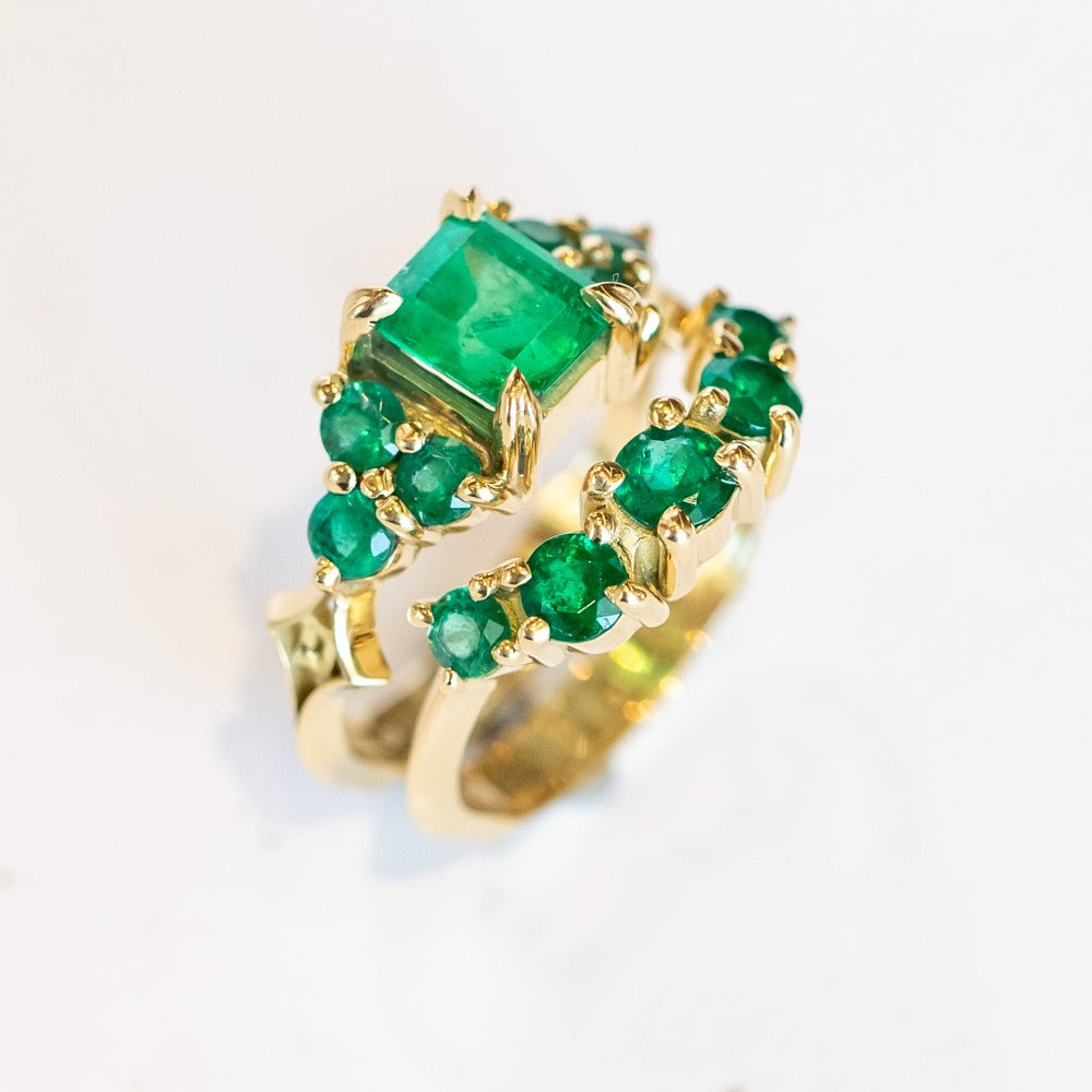 An emerald ring set of engagement ring and wedding band. Features an emerald cut emerald gemstone center flanked by six round emeralds, and a graduating size emerald band that contours to fit.