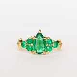 Yellow gold engagement ring featuring a pear-shaped emerald gemstone center stone flanked by six round emeralds, three on each side.