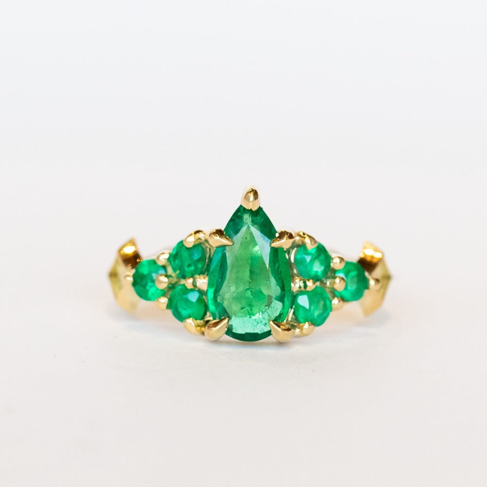 Yellow gold engagement ring featuring a pear-shaped emerald gemstone center stone flanked by six round emeralds, three on each side.
