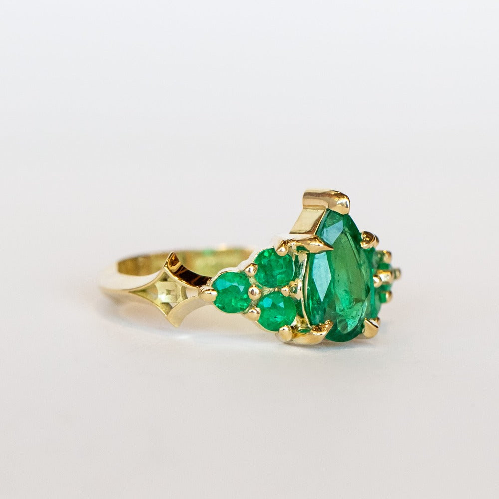Yellow gold engagement ring featuring a pear-shaped emerald gemstone center stone flanked by six round emeralds, three on each side.