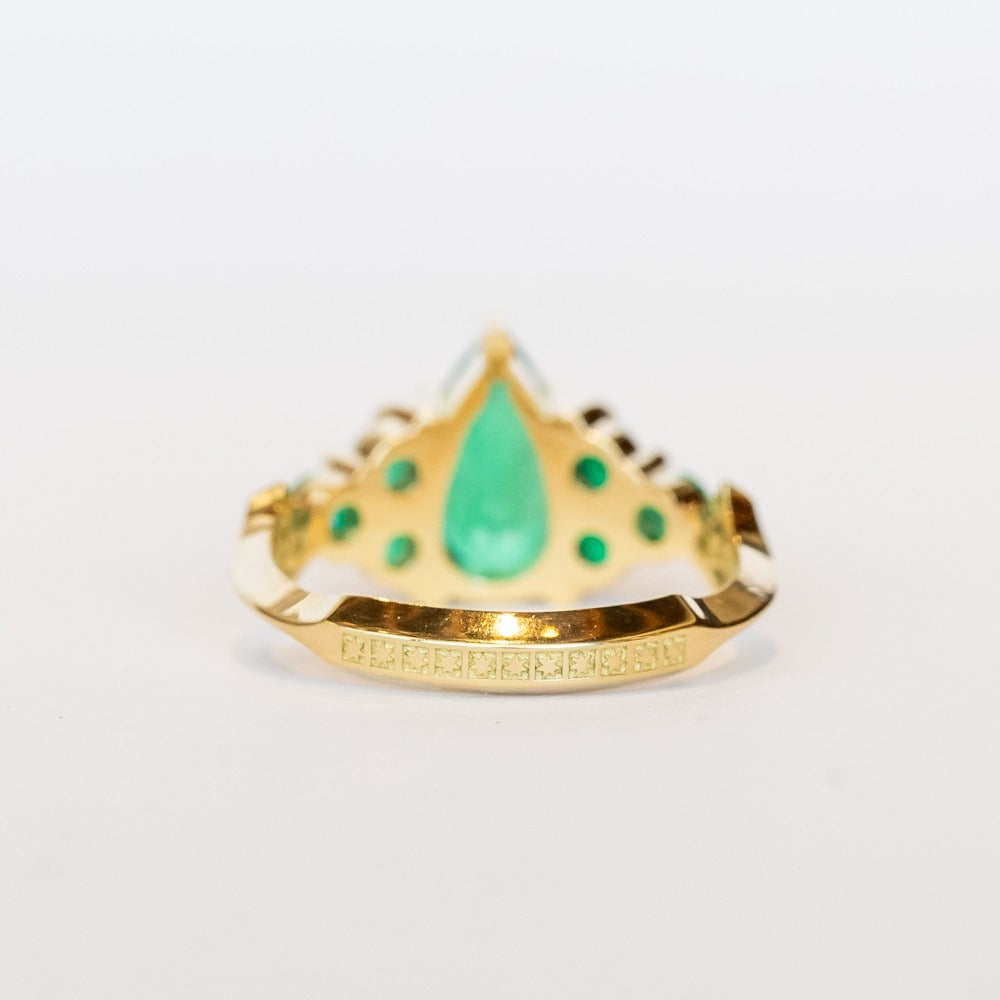 Yellow gold engagement ring featuring a pear-shaped emerald gemstone center stone flanked by six round emeralds, three on each side.