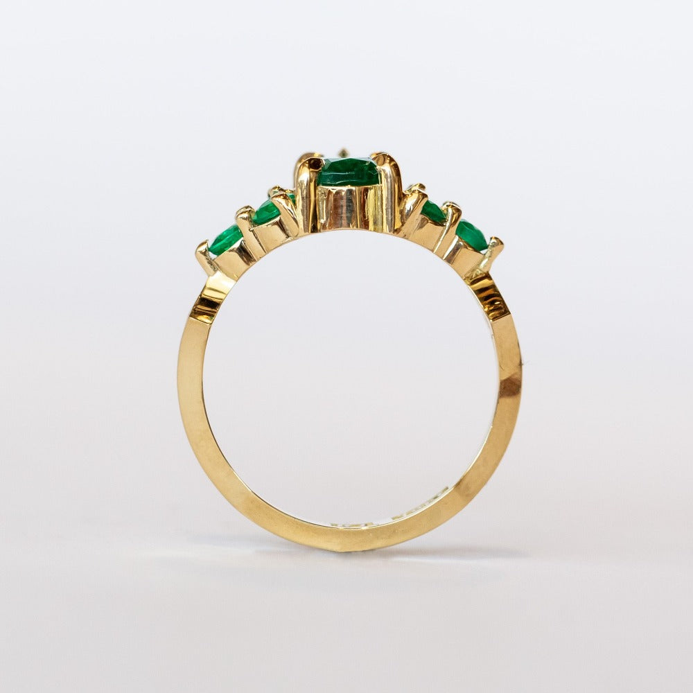 Yellow gold engagement ring featuring a pear-shaped emerald gemstone center stone flanked by six round emeralds, three on each side.
