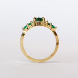 Yellow gold engagement ring featuring a pear-shaped emerald gemstone center stone flanked by six round emeralds, three on each side.