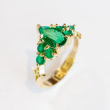 Yellow gold engagement ring featuring a pear-shaped emerald gemstone center stone flanked by six round emeralds, three on each side.