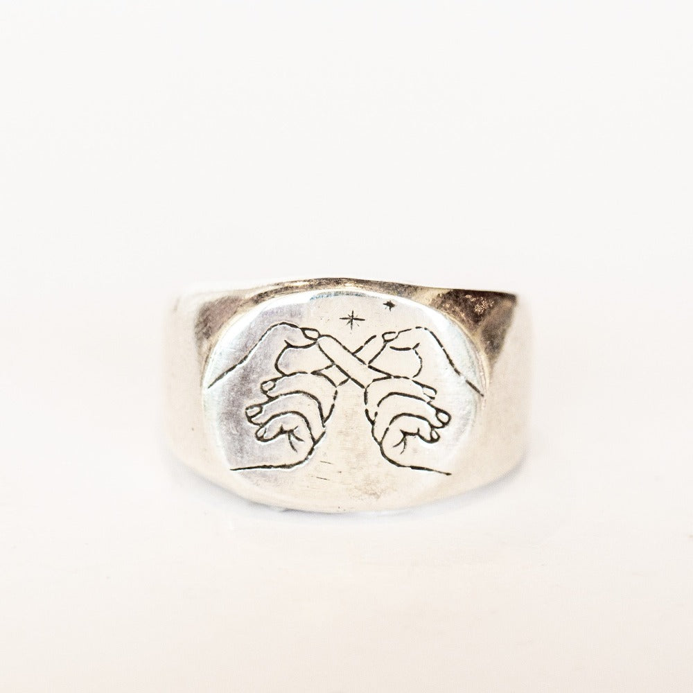 A chunky silver signet ring engraved with two hands making an infinity symbol and tiny stars.