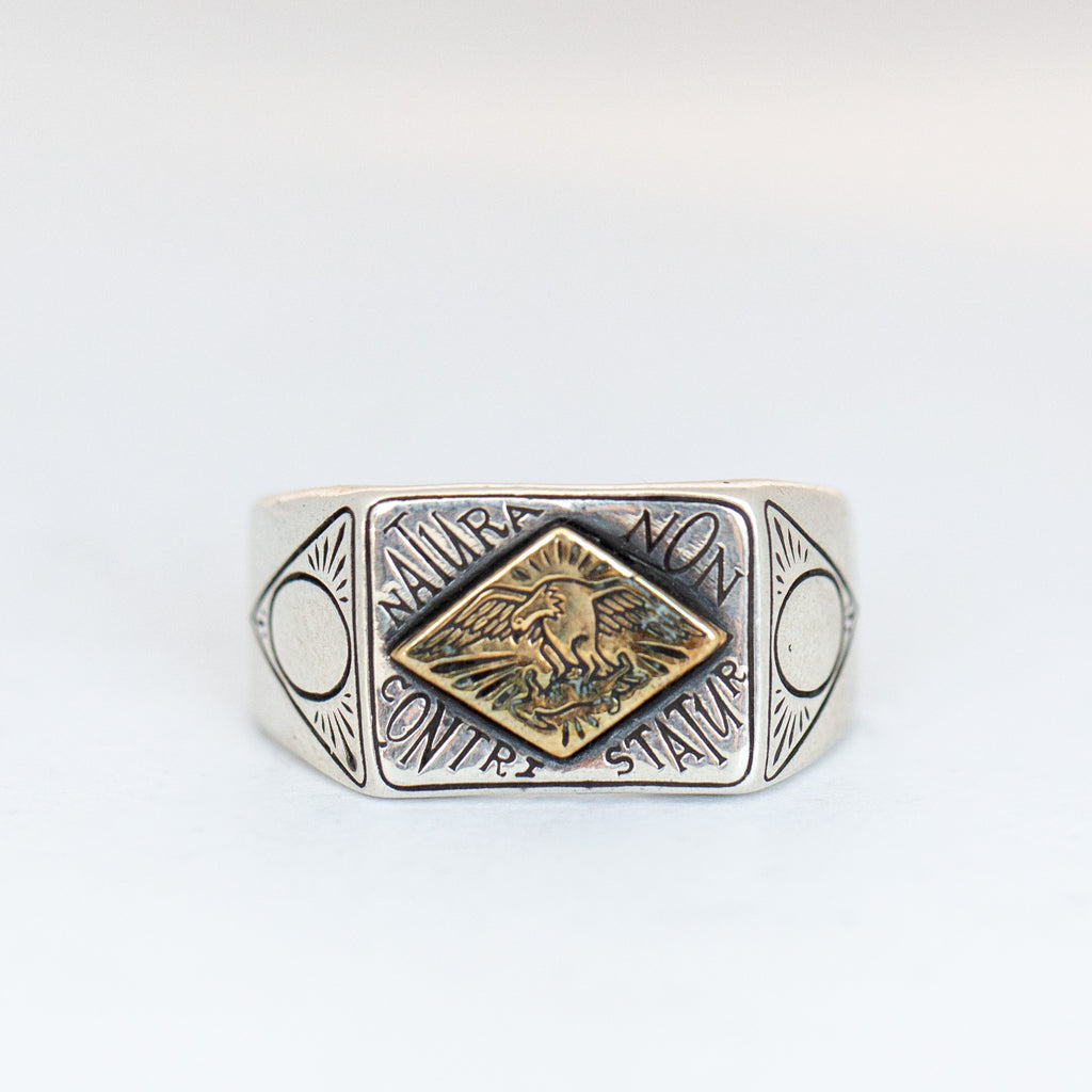 A chunky silver signet ring with diamond-shaped brass inlay. It is engraved with an eagle carrying away a rabbit, the ring reads 'Natura Non Constritatur', which translates from Latin to 'Nature Is Not Saddened'.