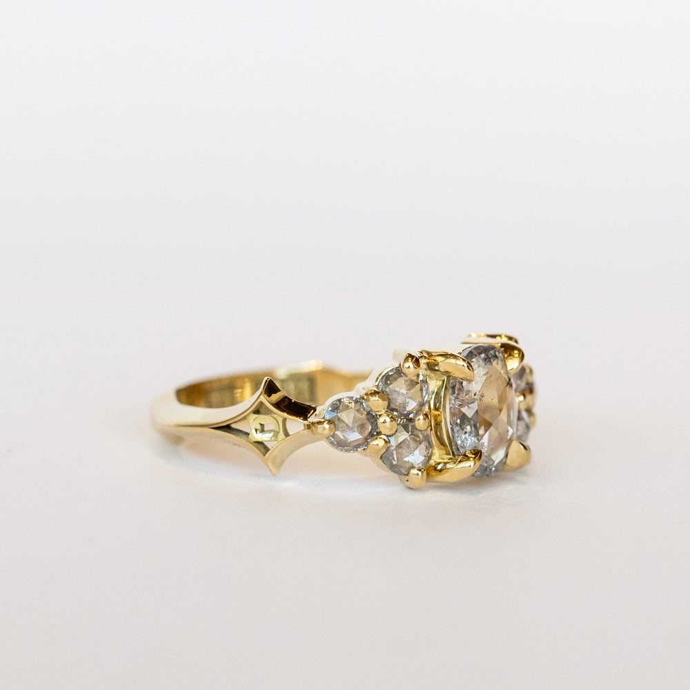A diamond engagement ring featuring salt and pepper diamonds, an oval at the center, flanked by clusters of three smaller diamonds on each side, set in 18 karat yellow gold.