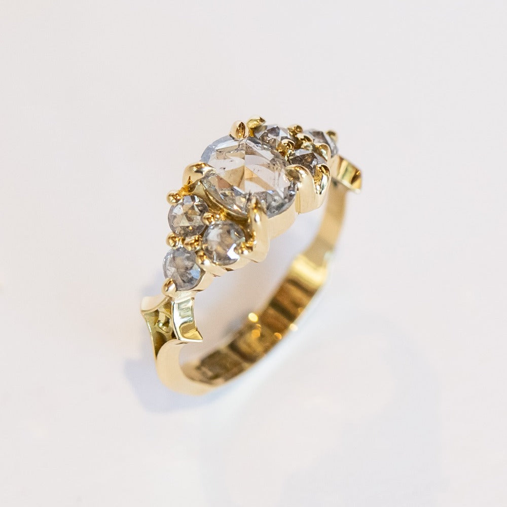 A diamond engagement ring featuring salt and pepper diamonds, an oval at the center, flanked by clusters of three smaller diamonds on each side, set in 18 karat yellow gold.