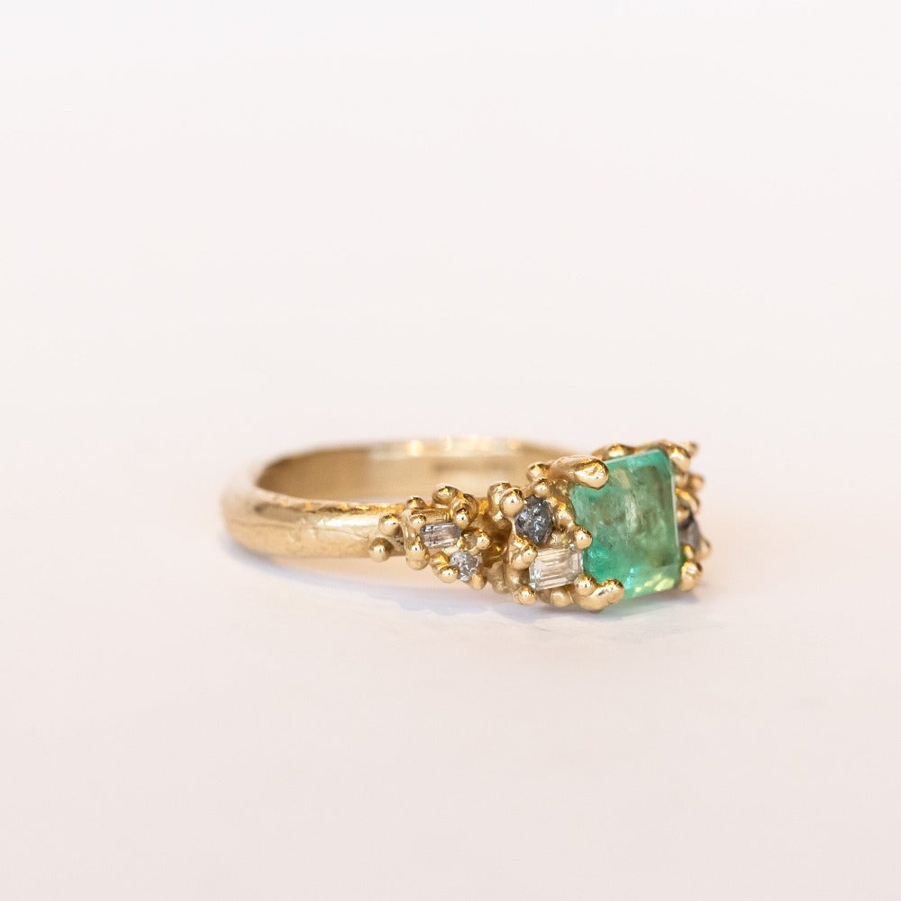 A yellow gold ring featuring an Asscher cut pale green emerald and clusters of diamonds to either side set in granulation.