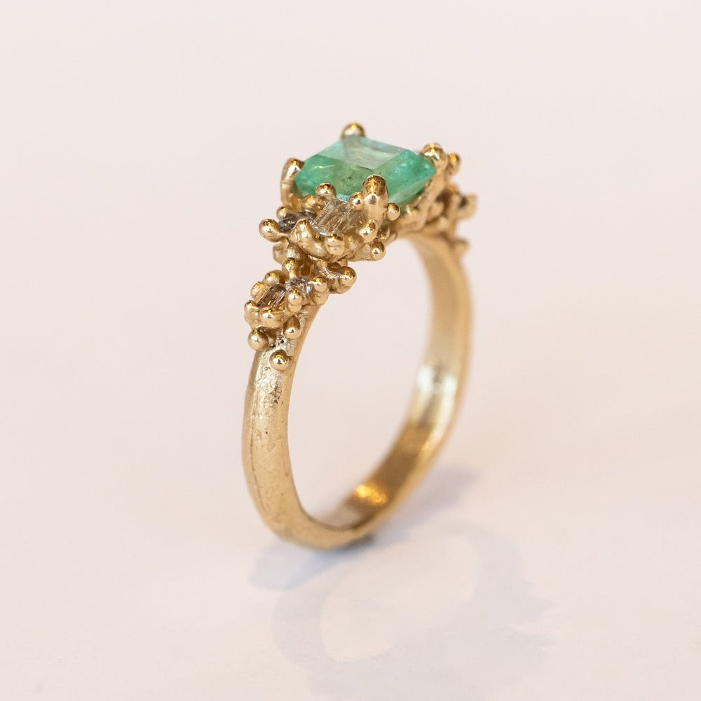 A yellow gold ring featuring an Asscher cut pale green emerald and clusters of diamonds to either side set in granulation.