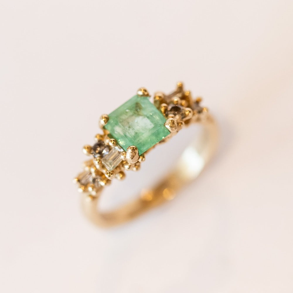 A yellow gold ring featuring an Asscher cut pale green emerald and clusters of diamonds to either side set in granulation.
