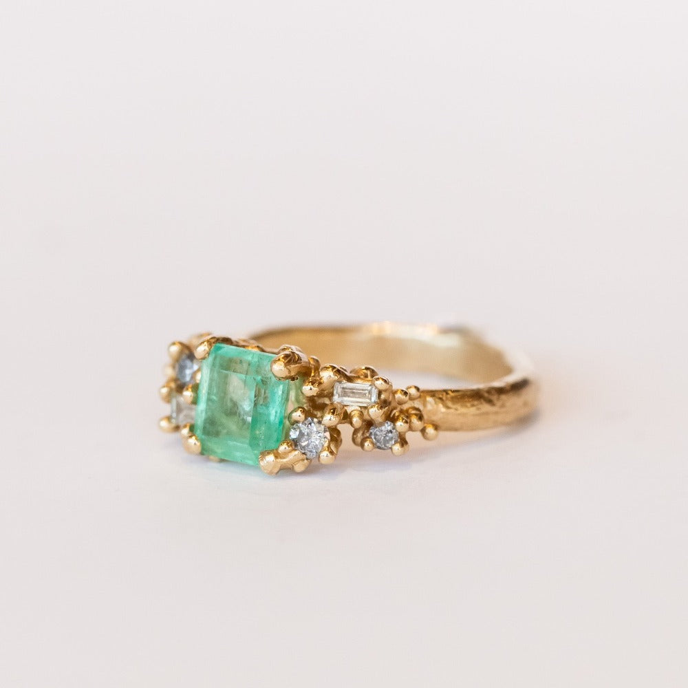 A yellow gold ring featuring an Asscher cut pale green emerald and clusters of diamonds to either side set in granulation.