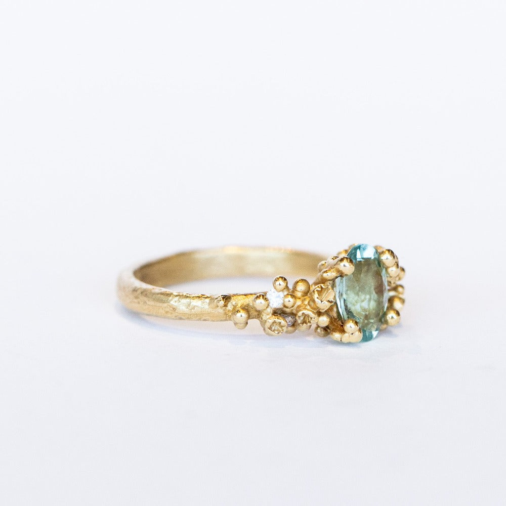 A pale blue-green oval tourmaline set in golden granules with diamond accents on a yellow gold ring.