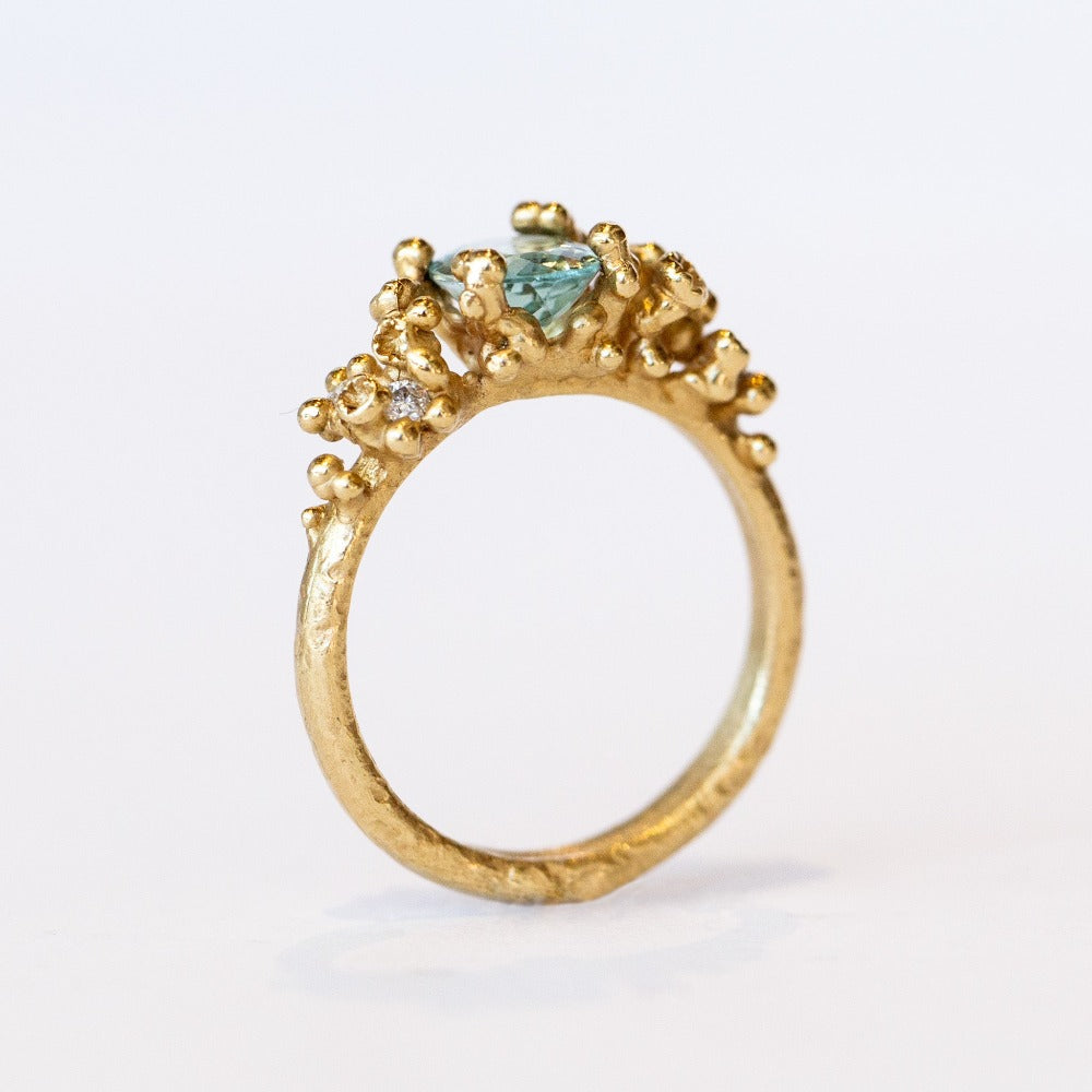 A pale blue-green oval tourmaline set in golden granules with diamond accents on a yellow gold ring.