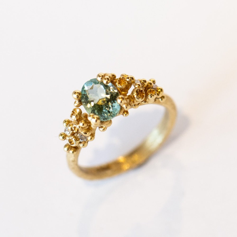 A pale blue-green oval tourmaline set in golden granules with diamond accents on a yellow gold ring.