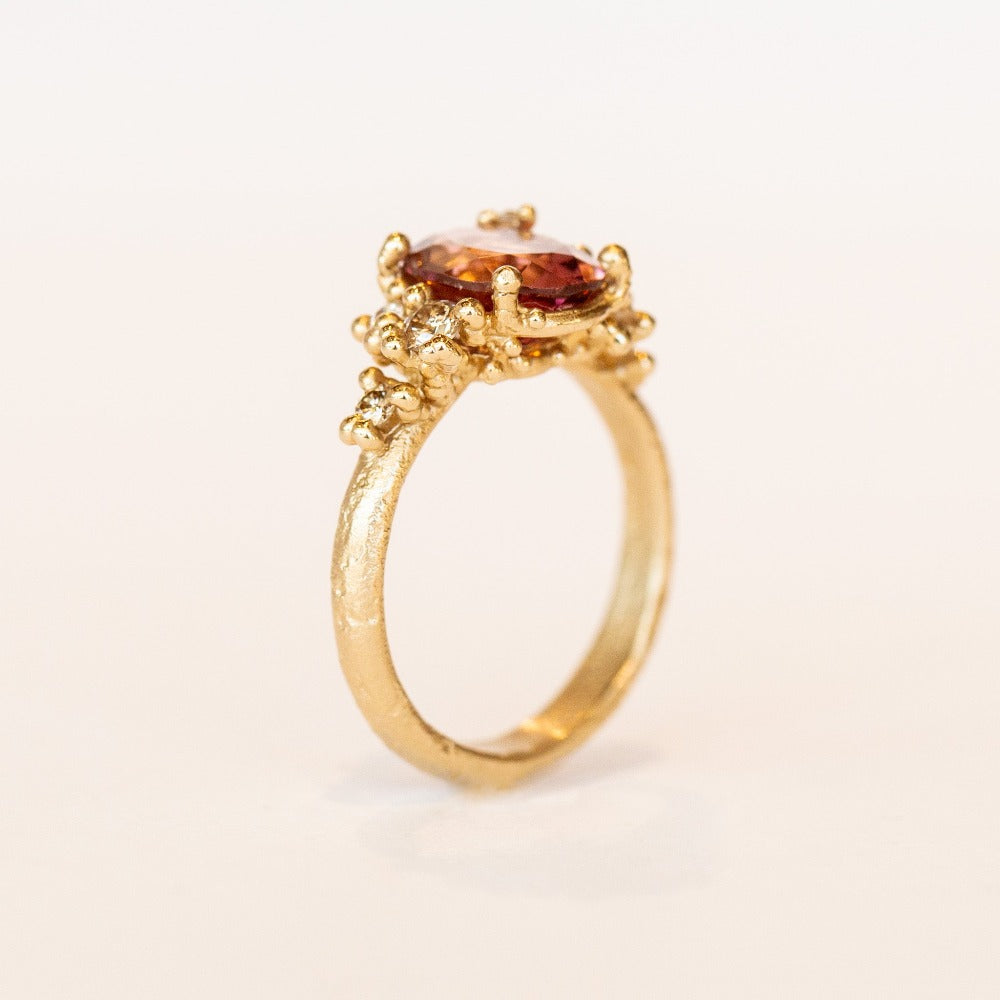 Oval peach-pink tourmaline set amongst&nbsp;golden granules and diamonds in yellow gold, on a gently textured band.