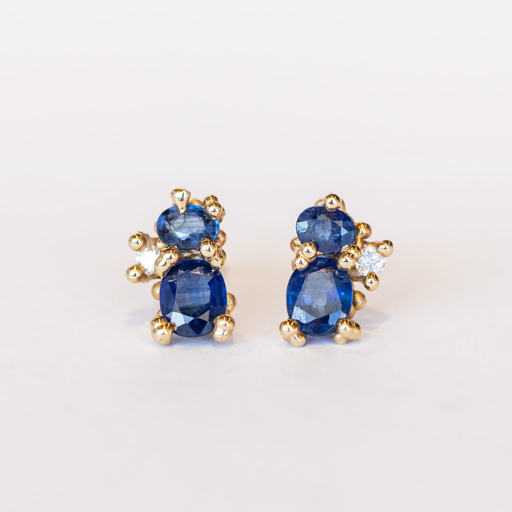 A pair of yellow gold stud earrings featuring two blue sapphires accented by tiny white diamonds and gold granules.