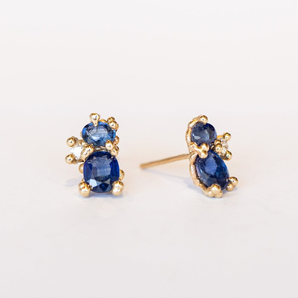 A pair of yellow gold stud earrings featuring two blue sapphires accented by tiny white diamonds and gold granules.
