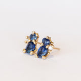 A pair of yellow gold stud earrings featuring two blue sapphires accented by tiny white diamonds and gold granules.