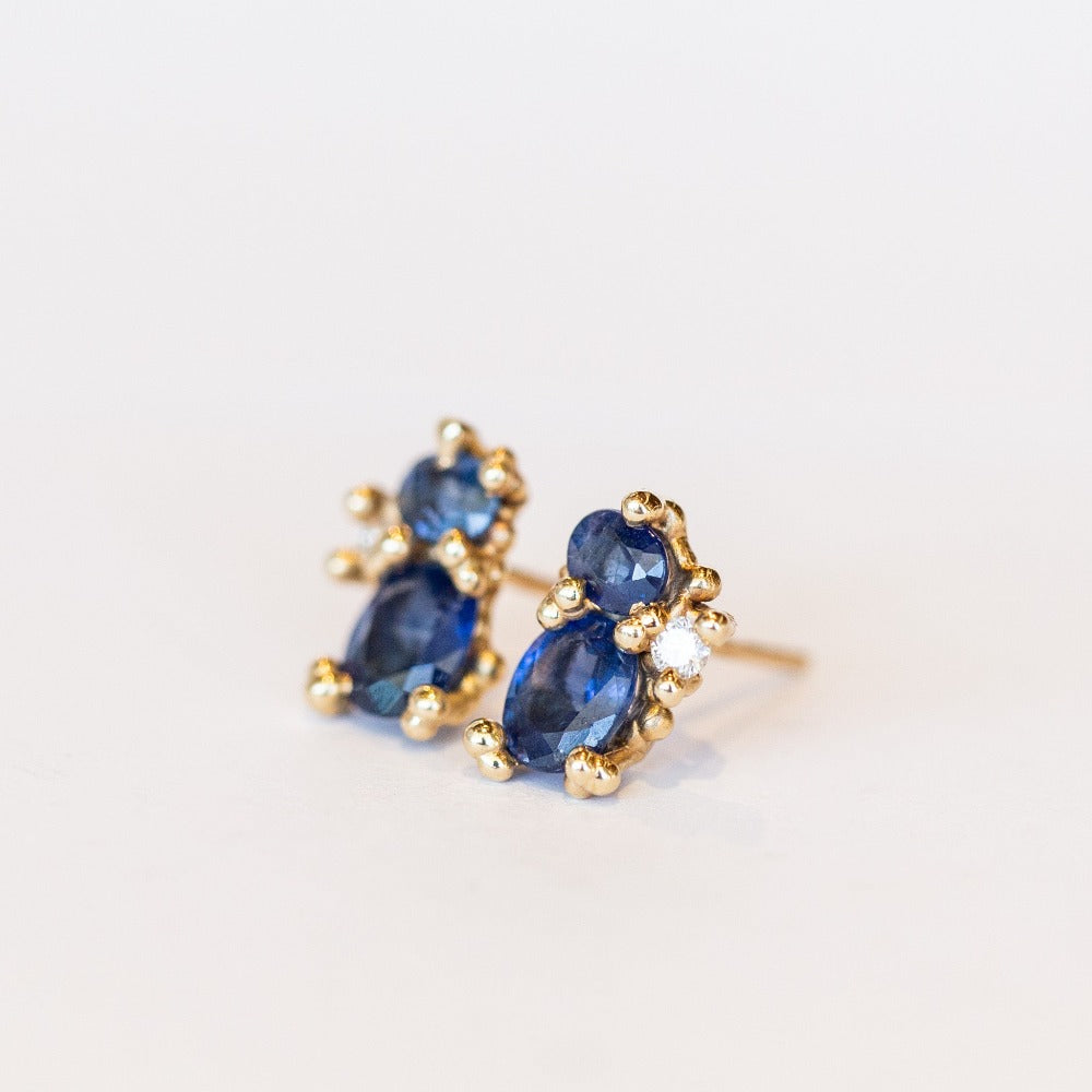 A pair of yellow gold stud earrings featuring two blue sapphires accented by tiny white diamonds and gold granules.