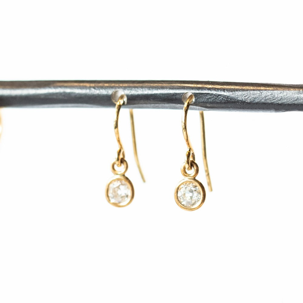 Diamond drop earrings, each featuring a single old european cut diamond, bezel set in gold, hanging from a gold ear hook wire.