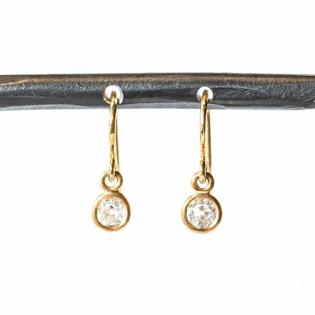 Diamond drop earrings, each featuring a single old european cut diamond, bezel set in gold, hanging from a gold ear hook wire.