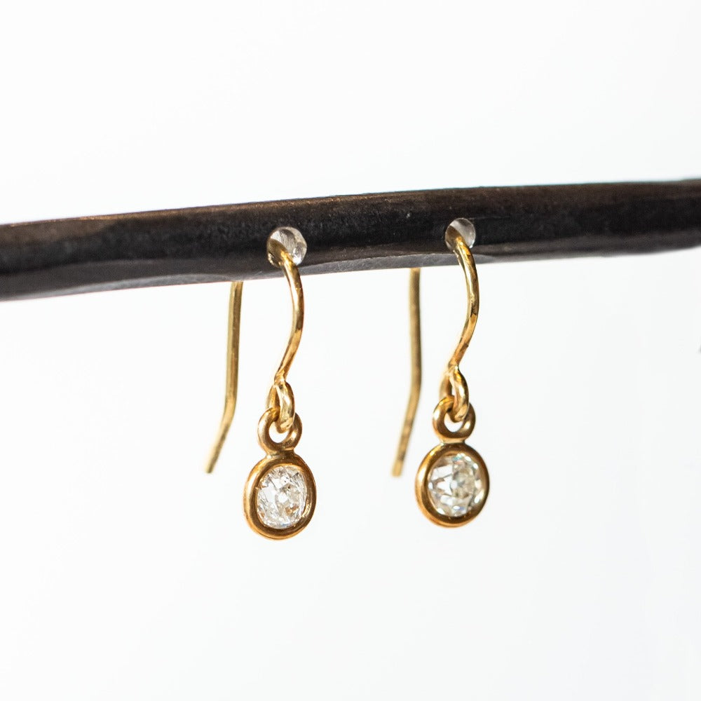 Diamond drop earrings, each featuring a single old european cut diamond, bezel set in gold, hanging from a gold ear hook wire.