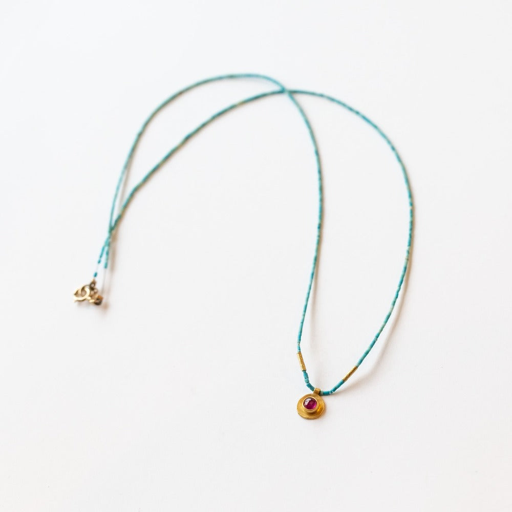 A thin, turquoise beaded necklace with a small ruby pendant set in yellow gold.
