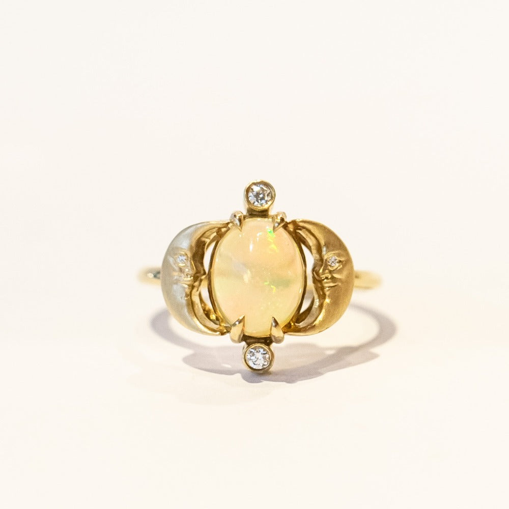 A yellow gold ring with an oval opal at its center, and facing moons on each side, accented by small diamonds.
