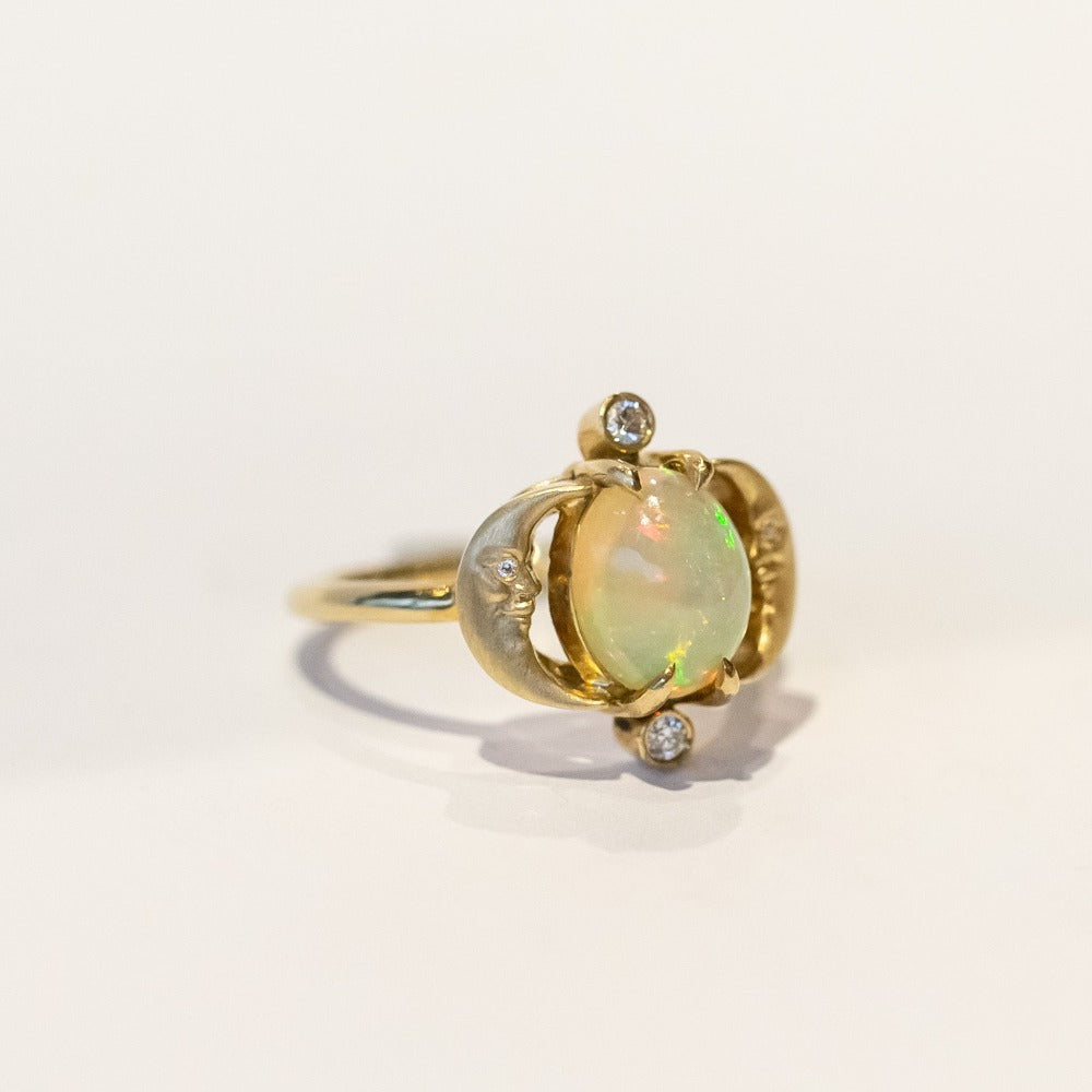 A yellow gold ring with an oval opal at its center, and facing moons on each side, accented by small diamonds.