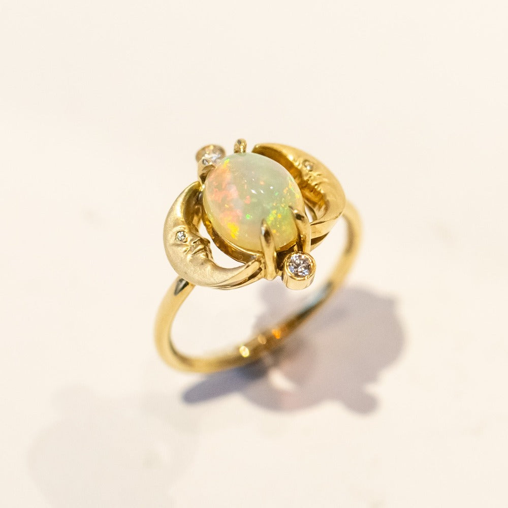 A yellow gold ring with an oval opal at its center, and facing moons on each side, accented by small diamonds.