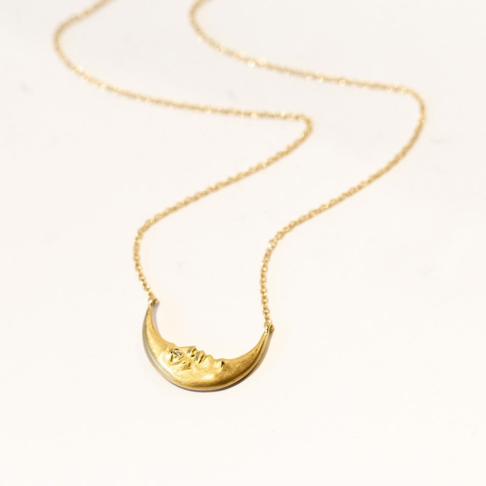 A horizontally-oriented crescent moon with face and diamond eye is secured at the center of a gold chain necklace.