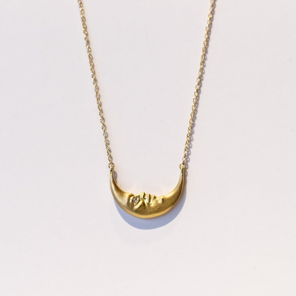 A horizontally-oriented crescent moon with face and diamond eye is secured at the center of a gold chain necklace.