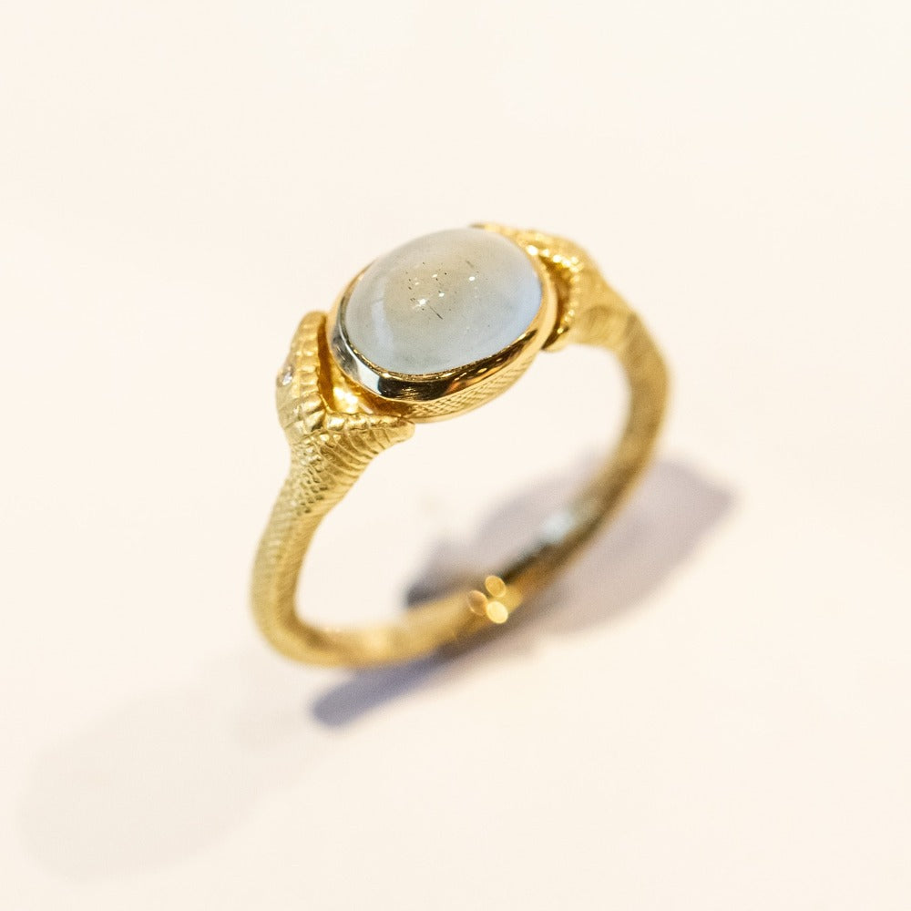 A carved yellow gold ring with an oval cabochon aquamarine at the center, held by open mouthed snakes on either side.