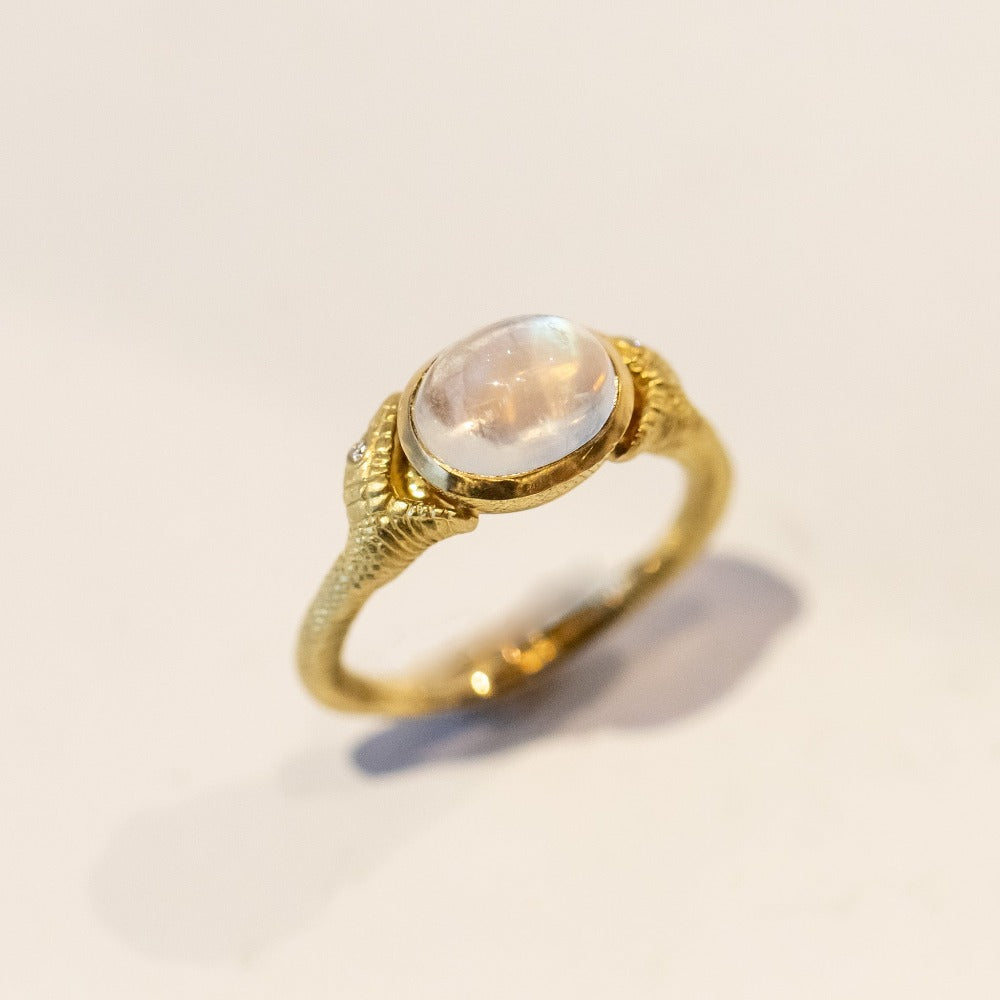 A carved yellow gold ring with an oval moonstone at the center, flanked by two open-mouthed snake heads with diamond eyes.