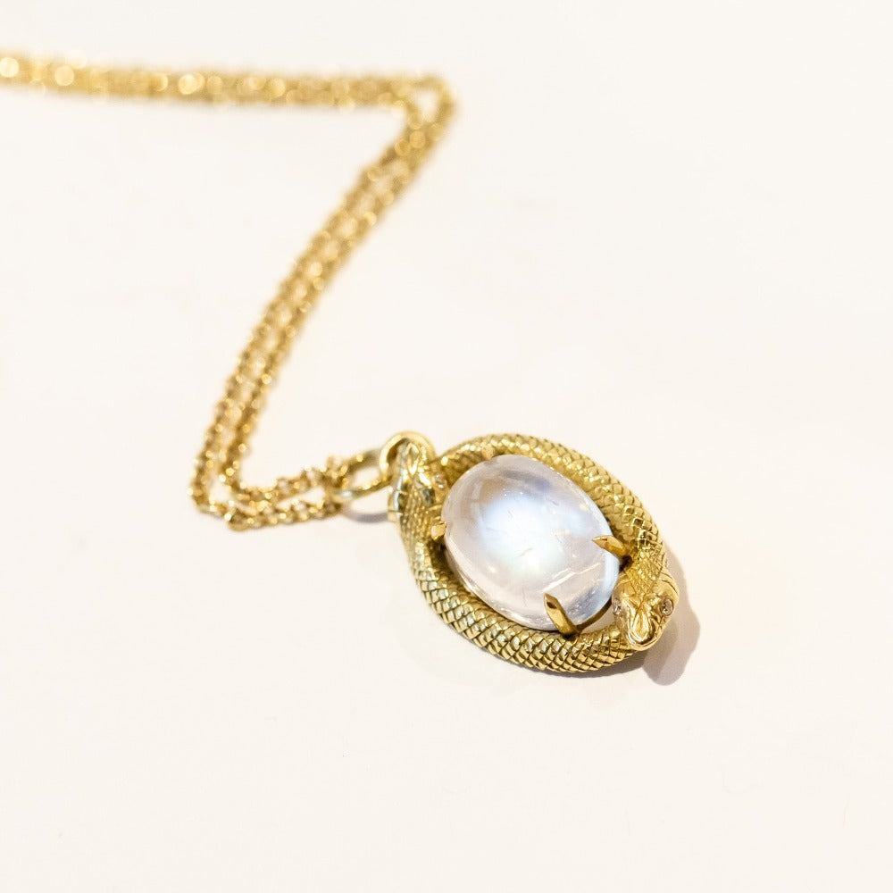 A pendant necklace featuring an oval moonstone at the center, encircled by two gold snakes in an ouroboros motif.