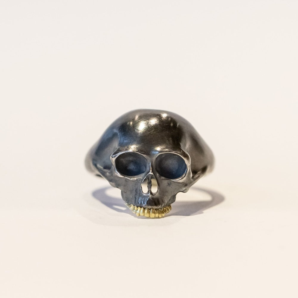A carved skull ring in blackened silver with gold teeth. Front view.