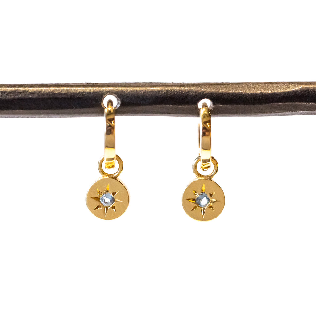 Dainty gold circle drops with a star-set aquamarine gemstone hang from small gold huggie hoop earrings.