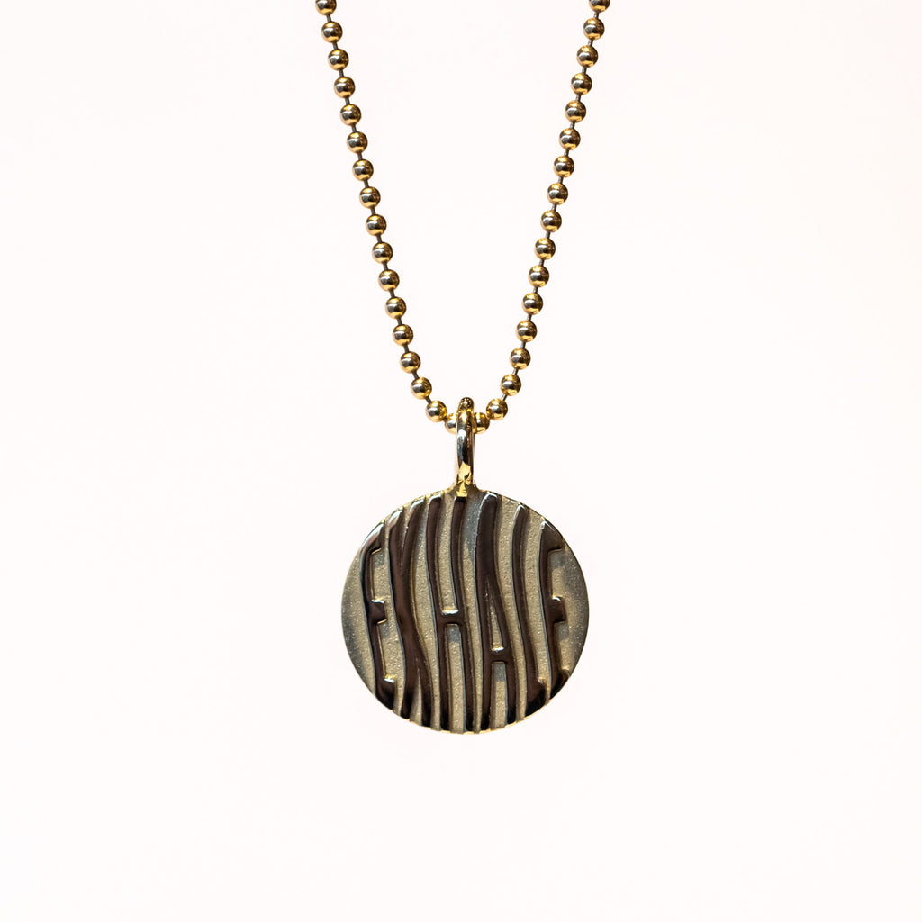 A round gold pendant with etched wavy letters spelling "exhale" hangs from a gold ball chain.