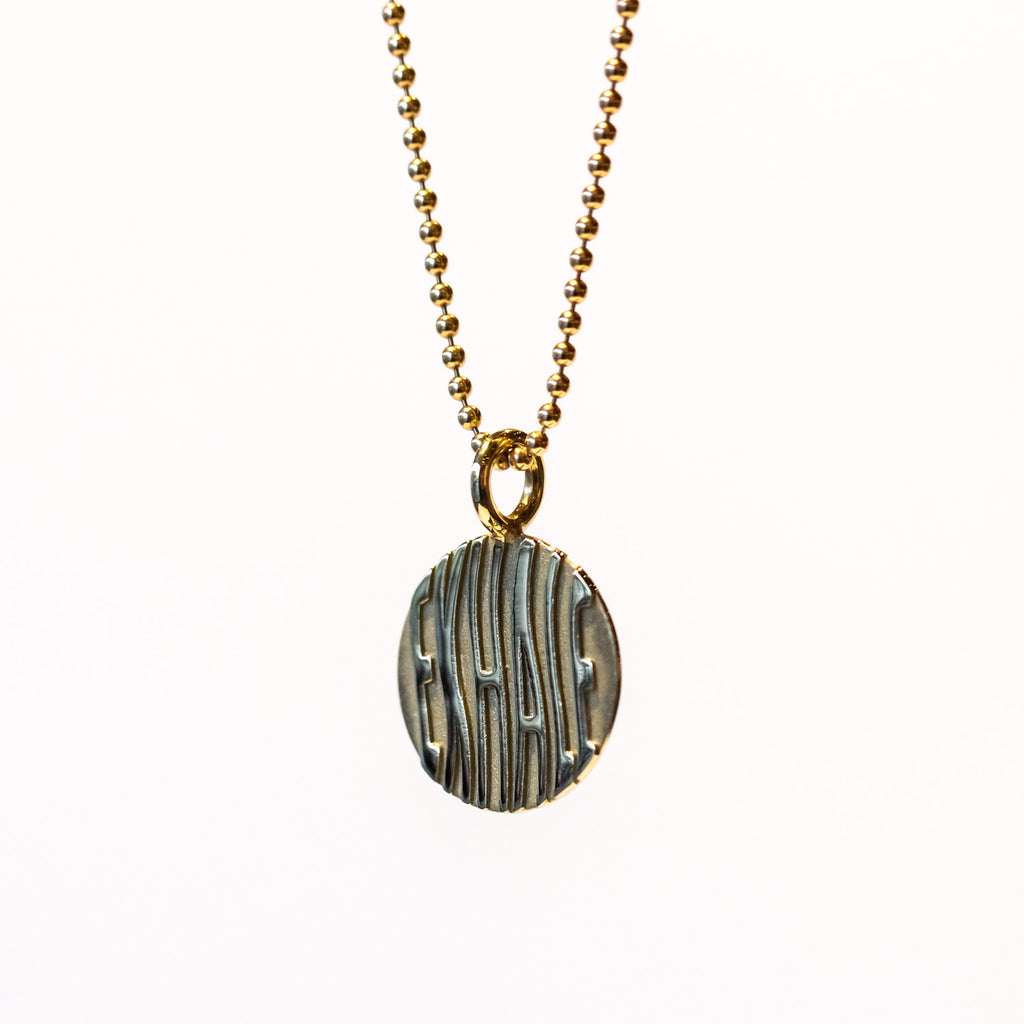 A round gold pendant with etched wavy letters spelling "exhale" hangs from a gold ball chain.