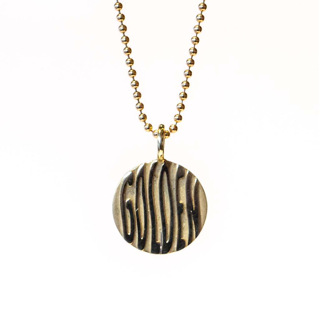 A round gold pendant featuring wavy letters spelling "golden" and suspended from a gold ball chain necklace.