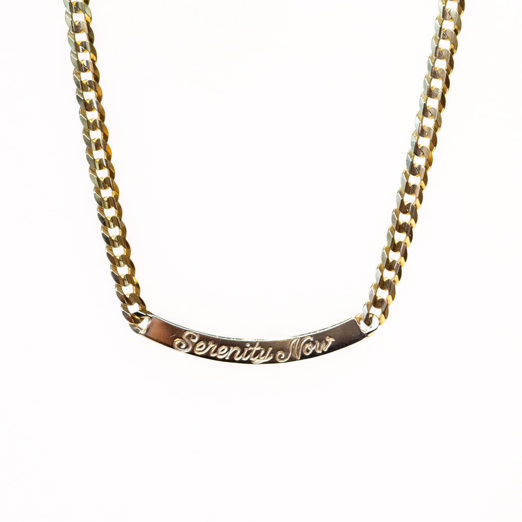 A gold curb chain necklace with a curved bar plate at the center, engraved in script that reads "serenity now."