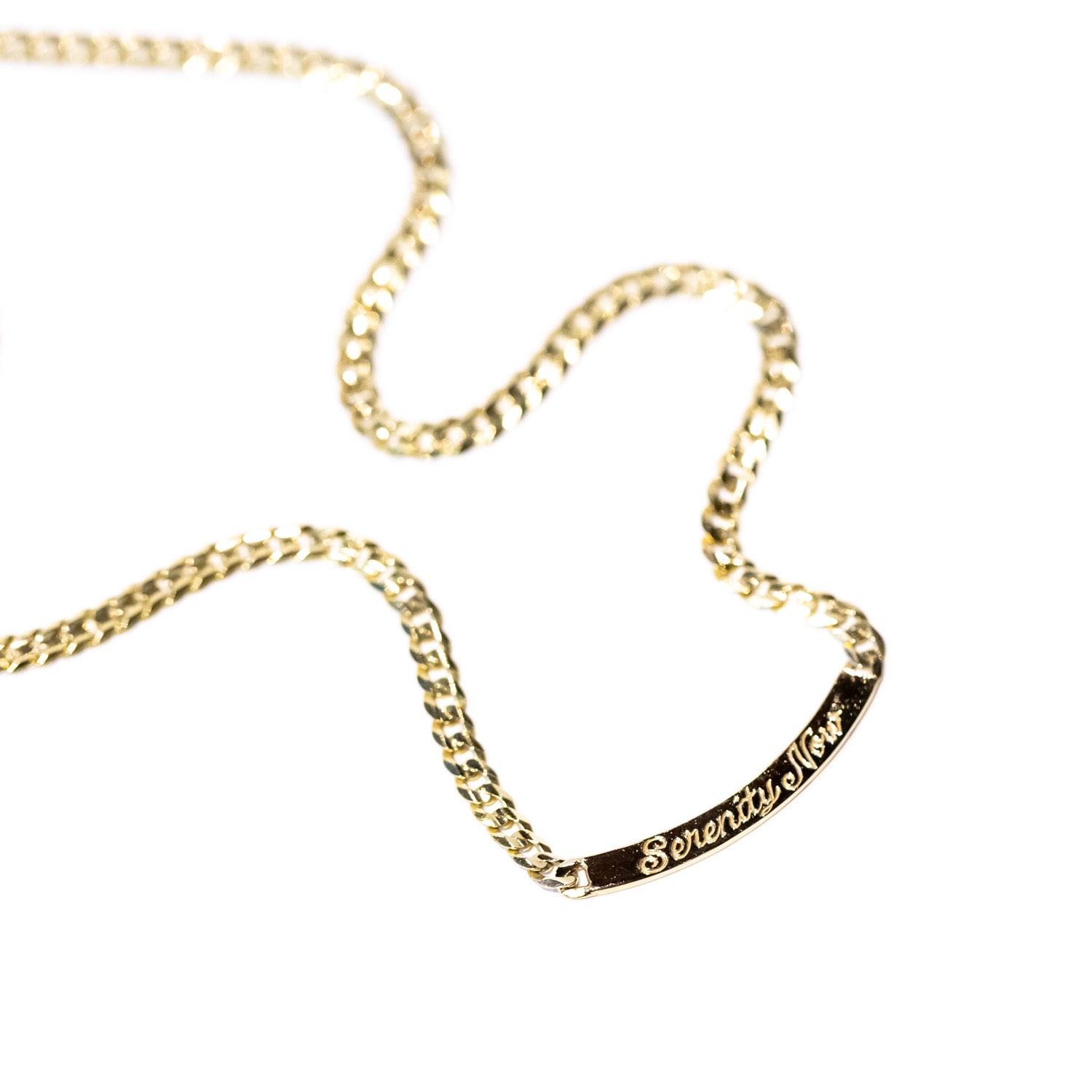 A gold curb chain necklace with a curved bar plate at the center, engraved in script that reads "serenity now."