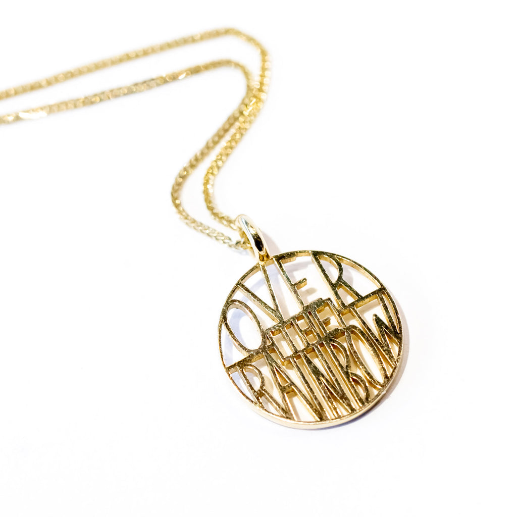 A circular gold pendant with thin lettering reading "over the rainbow".