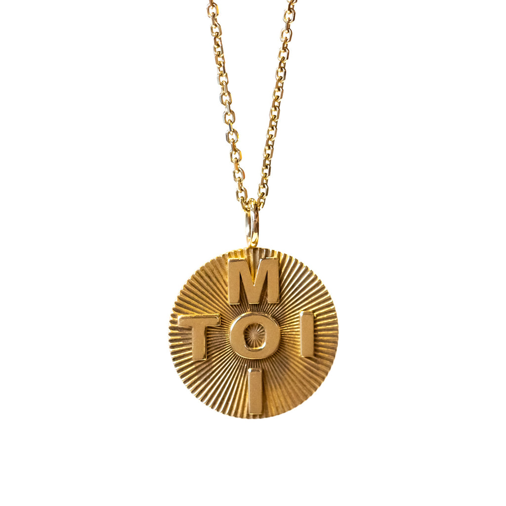 A round gold pendant with a carved starburst texture and the words "toi" and "moi" crossed in block letters through the middle. Hands from a simple gold chain necklace.